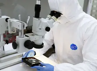 Technician in cleanroom suit examining a hard drive under a microscope for data recovery